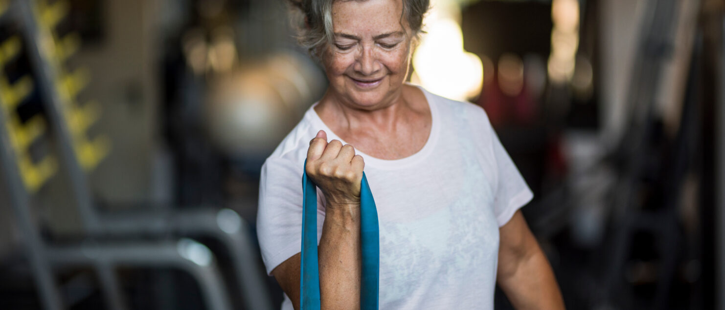 Ältere Frau führt im Fitnessstudio eine Armübung mit Gummiband durch.